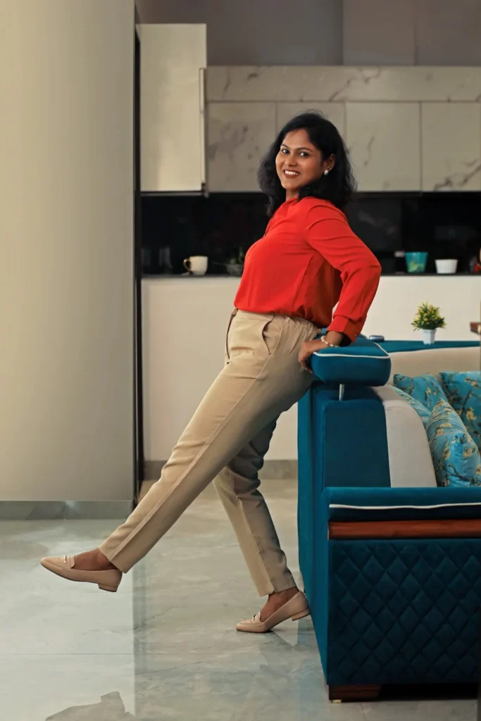 A confident professional digital marketing consultant in a red blouse and beige trousers smiling while leaning on a blue sofa.